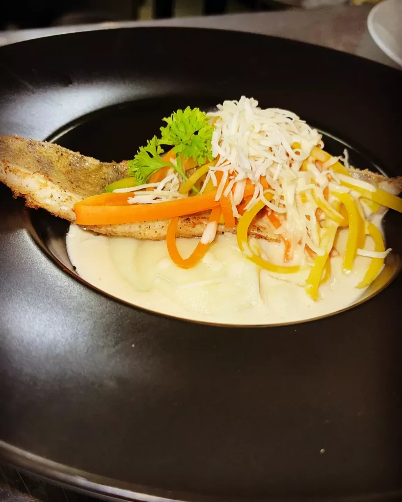 Gebratener Fischfilet auf cremiger Sauce mit Karotten- und Paprikastreifen, garniert mit Petersilie auf schwarzem Teller