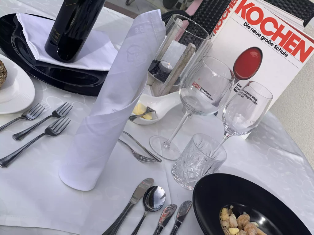 Eleganter gedeckter Tisch mit Weinflasche, weißen Servietten, Gläsern, Besteck und Kochbuch auf weißer Tischdecke