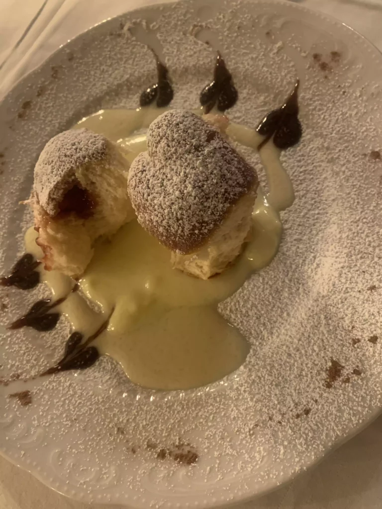 Zwei mit Puderzucker bestäubte Germknödel auf Vanillesoße mit Schokoladendekor auf weißem Teller in Naturhotel-Atmosphäre