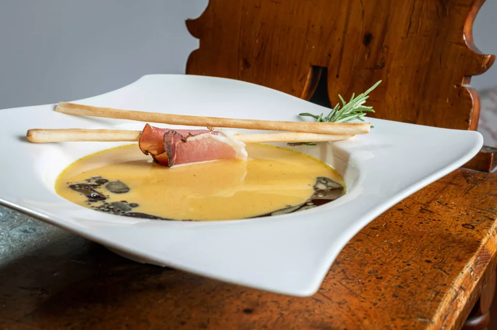 Cremige Suppe in weißem Teller mit zwei Grissini, einer Scheibe Schinken und Rosmarinzweig auf Holztisch vor Holzstuhl