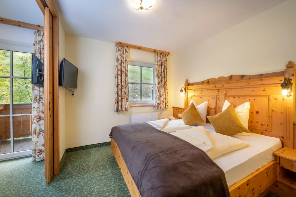 Gemütliches Hotelzimmer mit Holzmöbeln, Doppelbett, Fernseher, Fenster mit Blick ins Grüne und Zugang zum Balkon im
