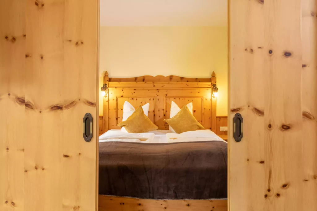 Blick durch offene Holztüren auf ein gemütliches Doppelbett mit Holzkopfteil und warmen Kissen in einem Naturhotelzimmer