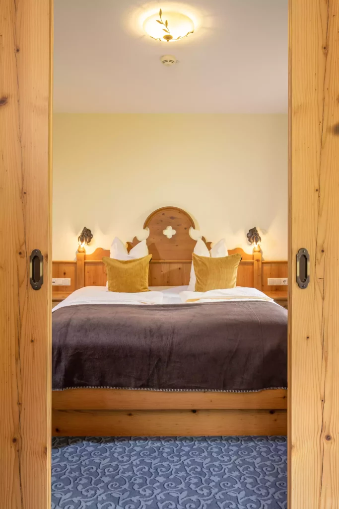 Hotelzimmer mit Doppelbett, Holzmöbeln, gelben Kissen und brauner Decke, Blick durch offene Holztüren