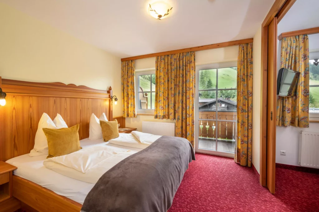 Gemütliches Hotelzimmer mit Doppelbett, gelben Vorhängen, rotem Teppich und Balkon mit Blick auf grüne Hügel im Großarltal