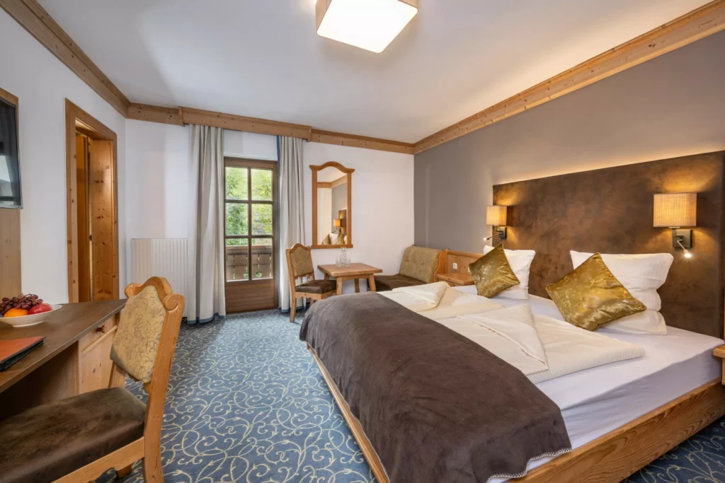 Gemütliches Hotelzimmer mit Doppelbett, goldenen Kissen, Holzmöbeln, Schreibtisch, Sitzbereich und Balkon mit Blick ins Grüne