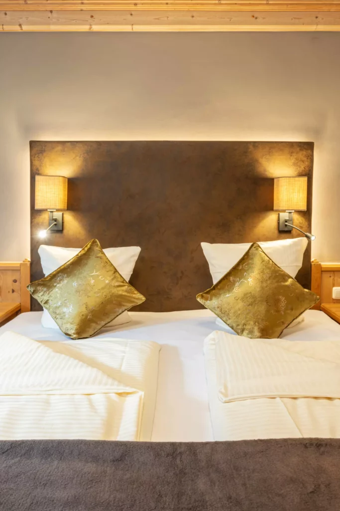 Doppelbett mit weißen Kissen und goldenen Zierkissen, braunes Kopfteil, zwei Wandlampen, gemütliches Naturhotelzimmer im