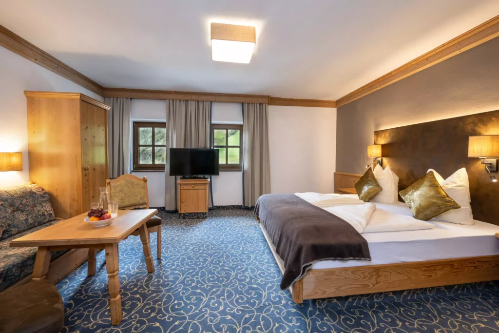 Gemütliches Hotelzimmer mit Doppelbett, Holzmöbeln, Fernseher, Tisch mit Stühlen und Blick auf grüne Natur durch Fenster in