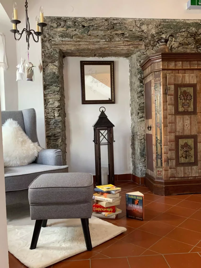 Gemütliche Sitzecke mit grauem Sessel, Hocker, Bücherstapel, antikem Schrank und Steinwand im Naturhotel Großarl