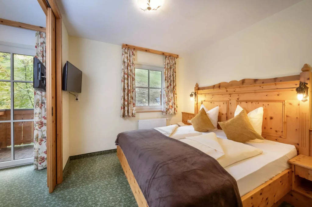 Gemütliches Hotelzimmer mit Doppelbett aus Holz, brauner Decke, zwei Fenstern mit gemusterten Vorhängen und Balkonblick auf