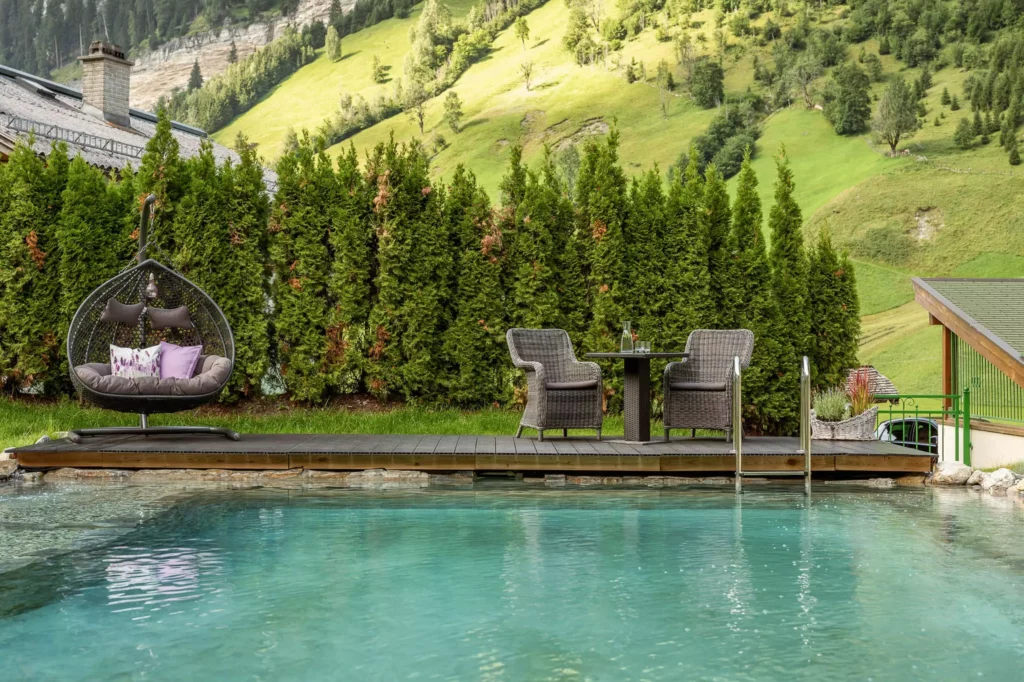 Außenpool mit Holzdeck, zwei Korbsesseln, Tisch mit Karaffe und Gläsern, Hängesessel und grüner Hecke im Naturhotel