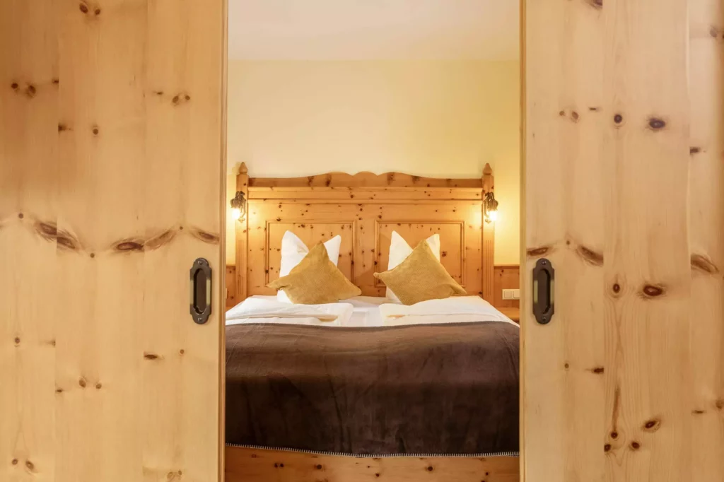 Blick durch offene Holztüren auf ein gemütliches Doppelbett mit Holzkopfteil und warmen Kissen im Naturhotel Hüttenwirt