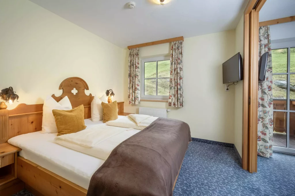 Gemütliches Hotelzimmer mit Doppelbett, Holzmöbeln, Fenster mit Blick auf grüne Wiese und Balkon im Naturhotel Hüttenwirt
