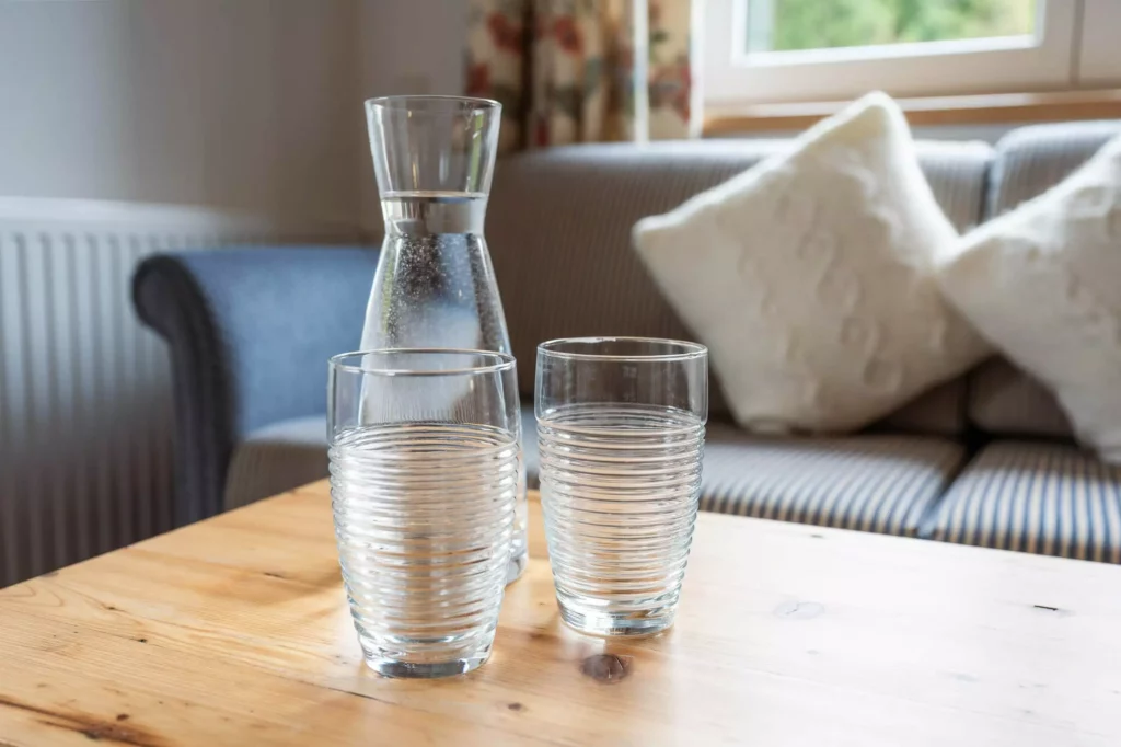 Zwei mit Wasser gefüllte Gläser und eine Karaffe auf einem Holztisch vor einem Sofa mit Kissen im Naturhotel Hüttenwirt