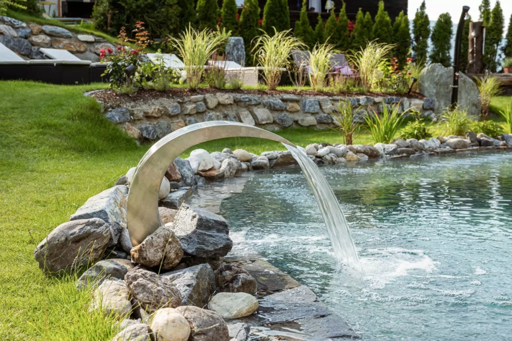 Wasserfontäne aus gebogenem Metallrohr in Naturpool mit Steinen und Liegestühlen im Garten des Naturhotel Hüttenwirt Großarl