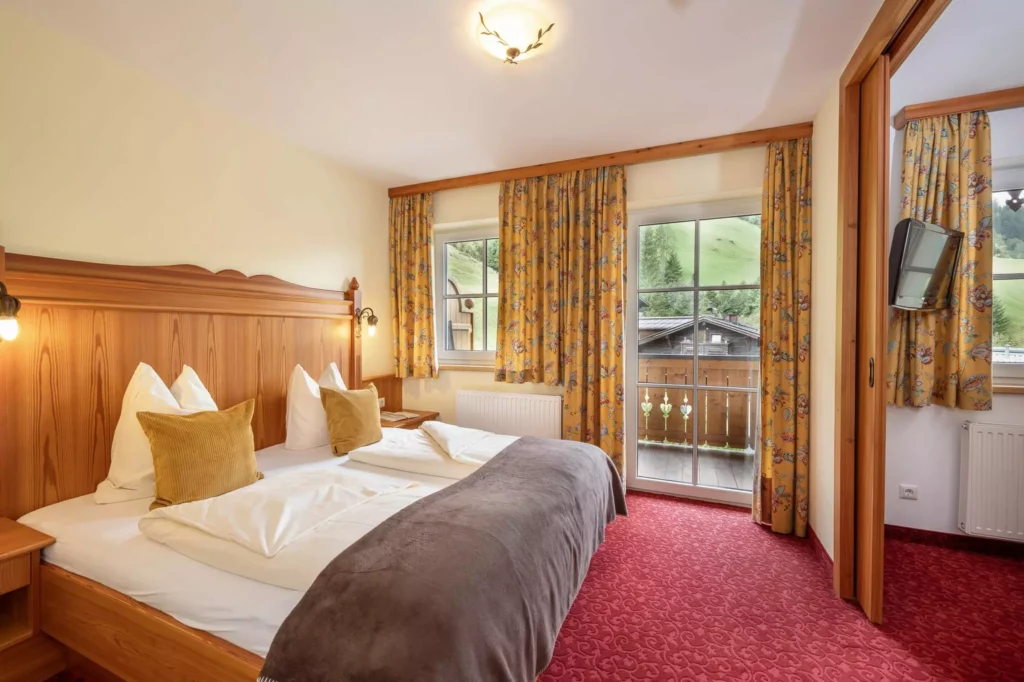 Hotelzimmer mit Doppelbett, Holzmöbeln, gelben Vorhängen, rotem Teppich und Balkon mit Bergblick im Naturhotel Hüttenwirt