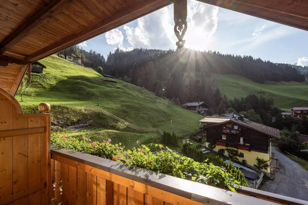 Blick von einem Holz-Balkon mit Blumenkasten auf grüne Hügel, Wald und traditionelle Häuser im Großarltal bei Sonnenschein