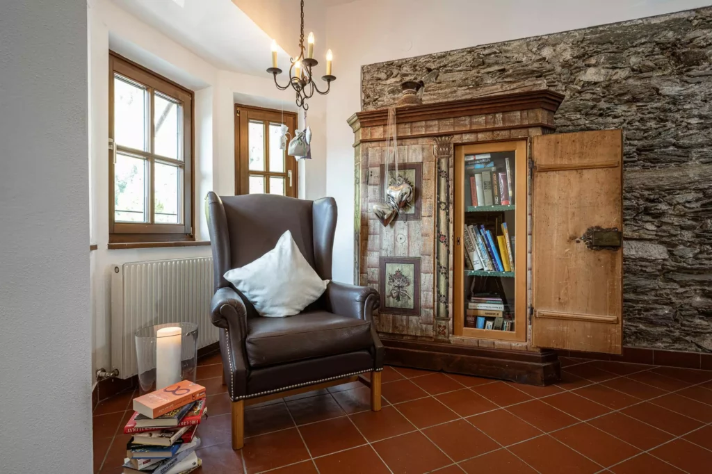 Gemütliche Leseecke mit braunem Ledersessel, weißem Kissen, Kerze auf Bücherstapel und antikem Bücherschrank im Naturhotel