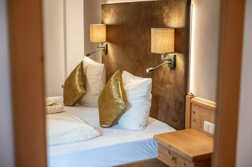 Gemütliches Hotelzimmer mit Doppelbett, goldfarbenen Kissen, Wandlampen und Nachttisch aus Holz im Naturhotel Hüttenwirt