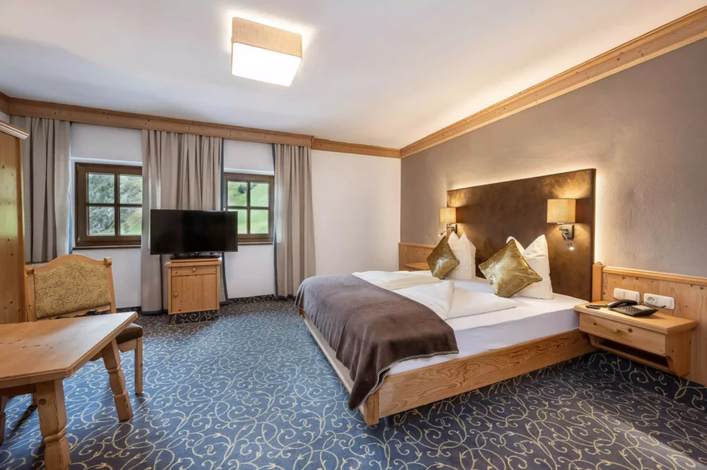 Gemütliches Hotelzimmer mit Doppelbett, Holzmöbeln, Teppichboden und Fernseher vor zwei Fenstern mit Blick auf grüne Berge