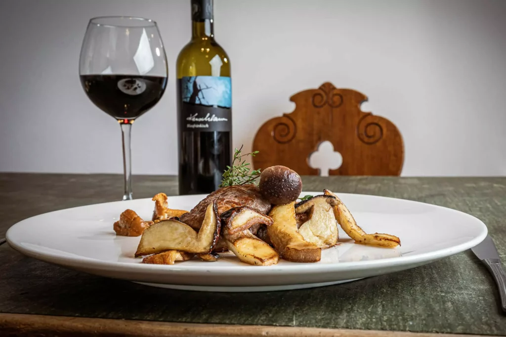 Gebratenes Pilzgericht auf weißem Teller mit Glas Rotwein und Weinflasche auf rustikalem Holztisch im Naturhotel Hüttenwirt