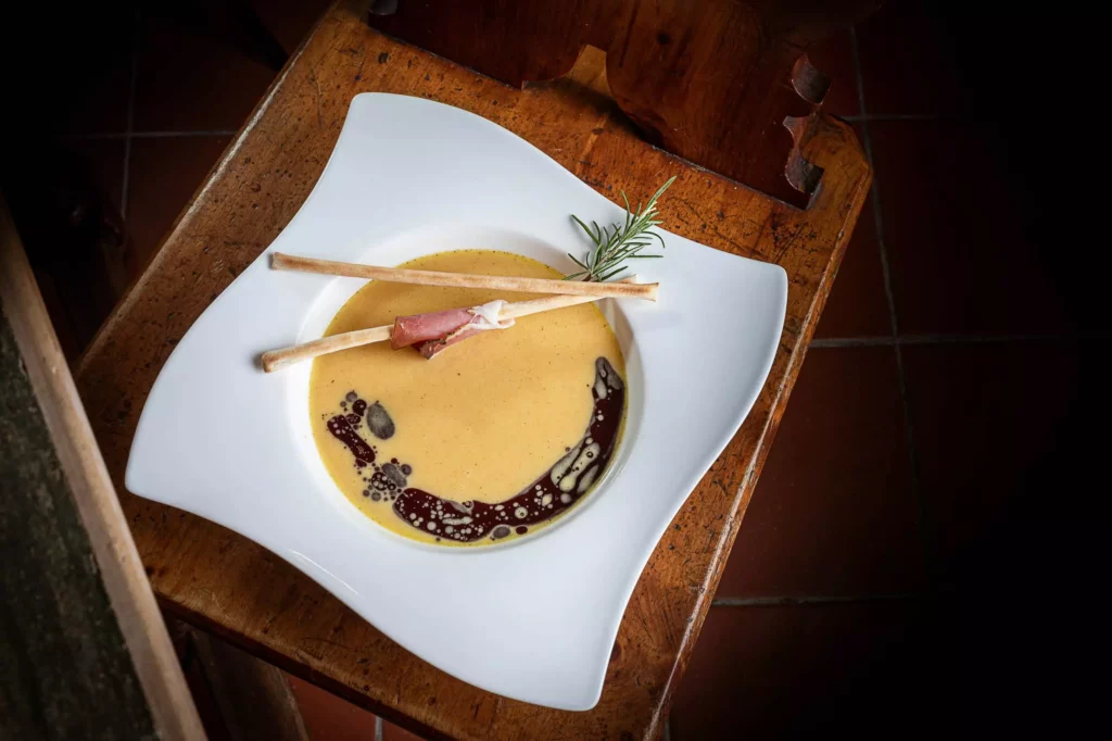 Cremige Suppe mit dunklem Öl, zwei dünnen Brotstangen, einer Scheibe Schinken und Rosmarin auf weißem Teller auf Holzstuhl