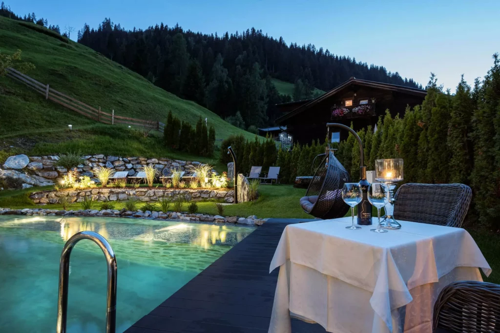 Abendliche Poollandschaft mit gedecktem Tisch, Weinflasche, Gläsern und gemütlicher Sitzgelegenheit im Naturhotel Hüttenwirt