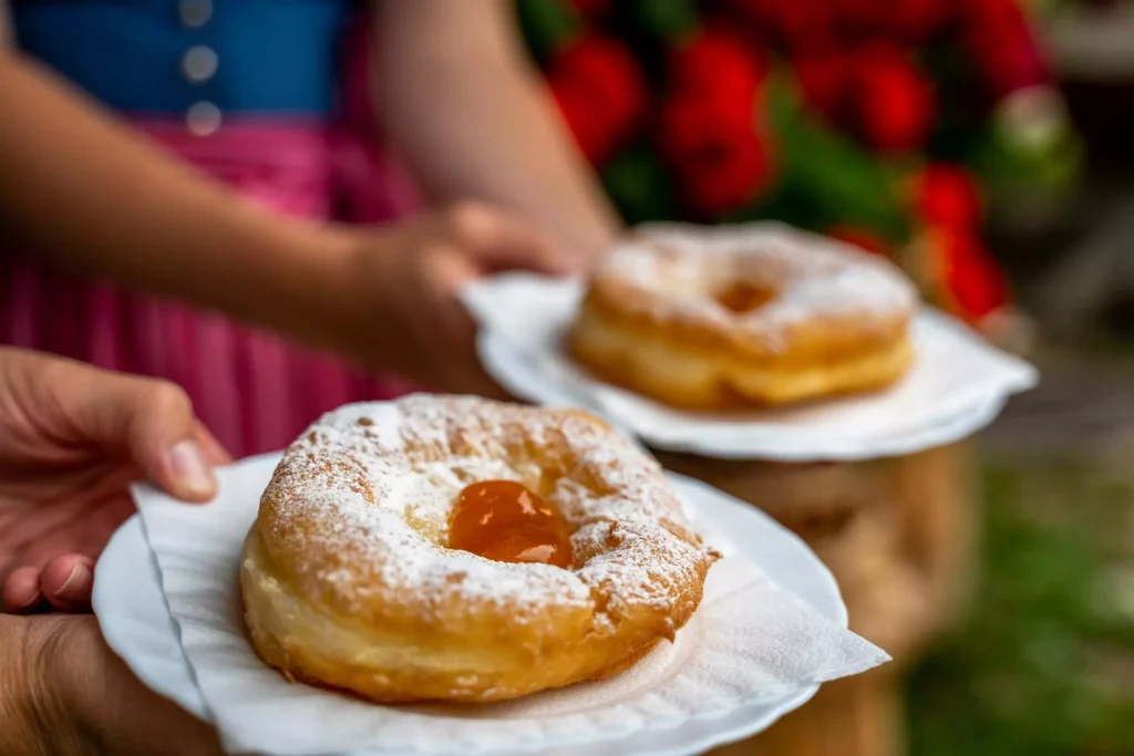 Zwei Hände halten Teller mit Staubzucker bestäubten Krapfen mit Marillenmarmelade, unscharfer Hintergrund mit Blumen.