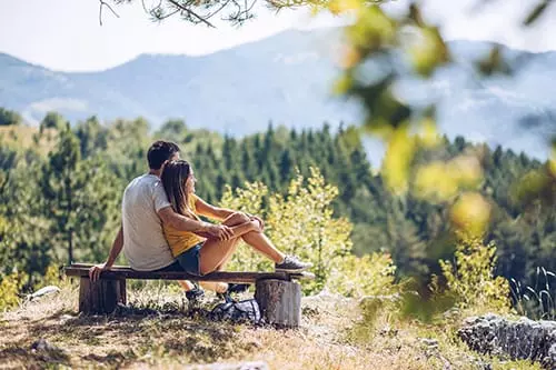Paar sitzt auf Holzbank in der Natur mit Blick auf bewaldete Berge im Großarltal, entspannte Atmosphäre im Sommer.