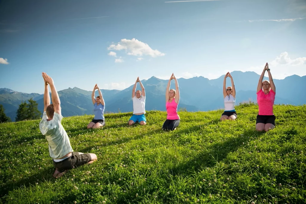 Sechs Personen praktizieren Yoga kniend auf einer grünen Wiese vor Bergpanorama bei Sonnenschein im Großarltal.